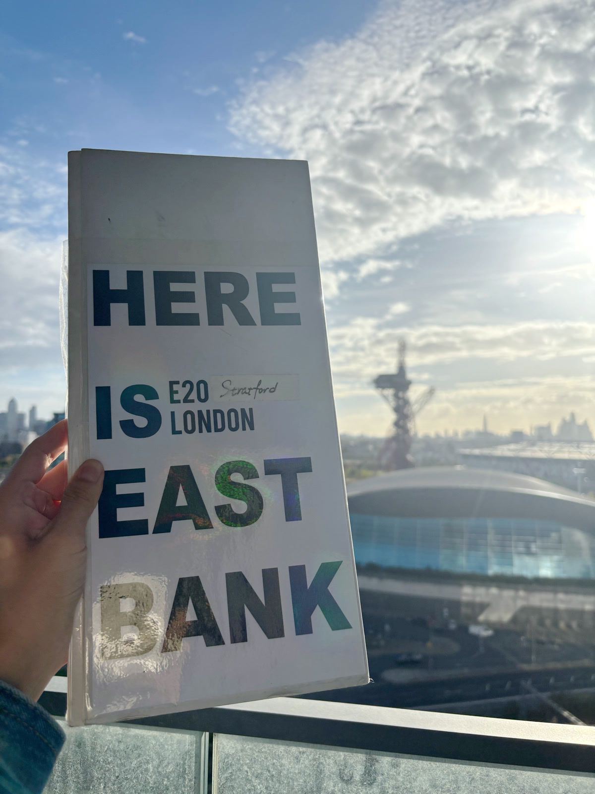 Here is East Bank — the book dummy held up against a blue sky, with the Olympic Park and the ArcelorMittal Orbit in the background