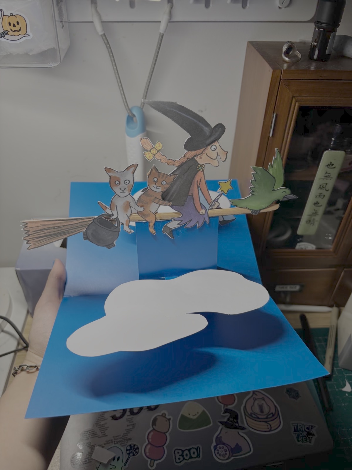 The same pop-up card opened on a desk, seen from closer: the witch, cat, cauldron and crow standing up from the blue card.