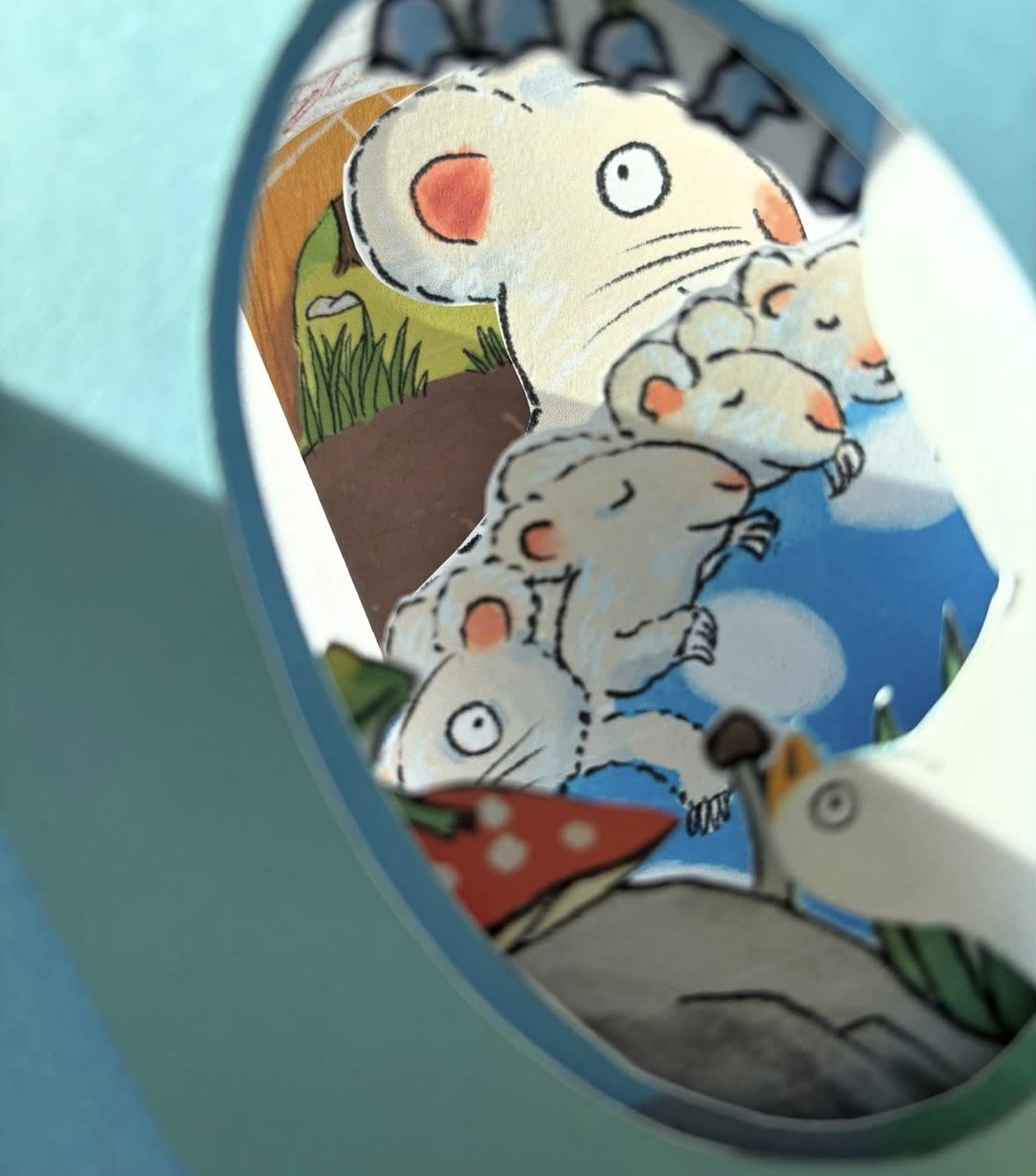 A close view through a round window of a pop-up card: a family of mice among mushrooms and grass