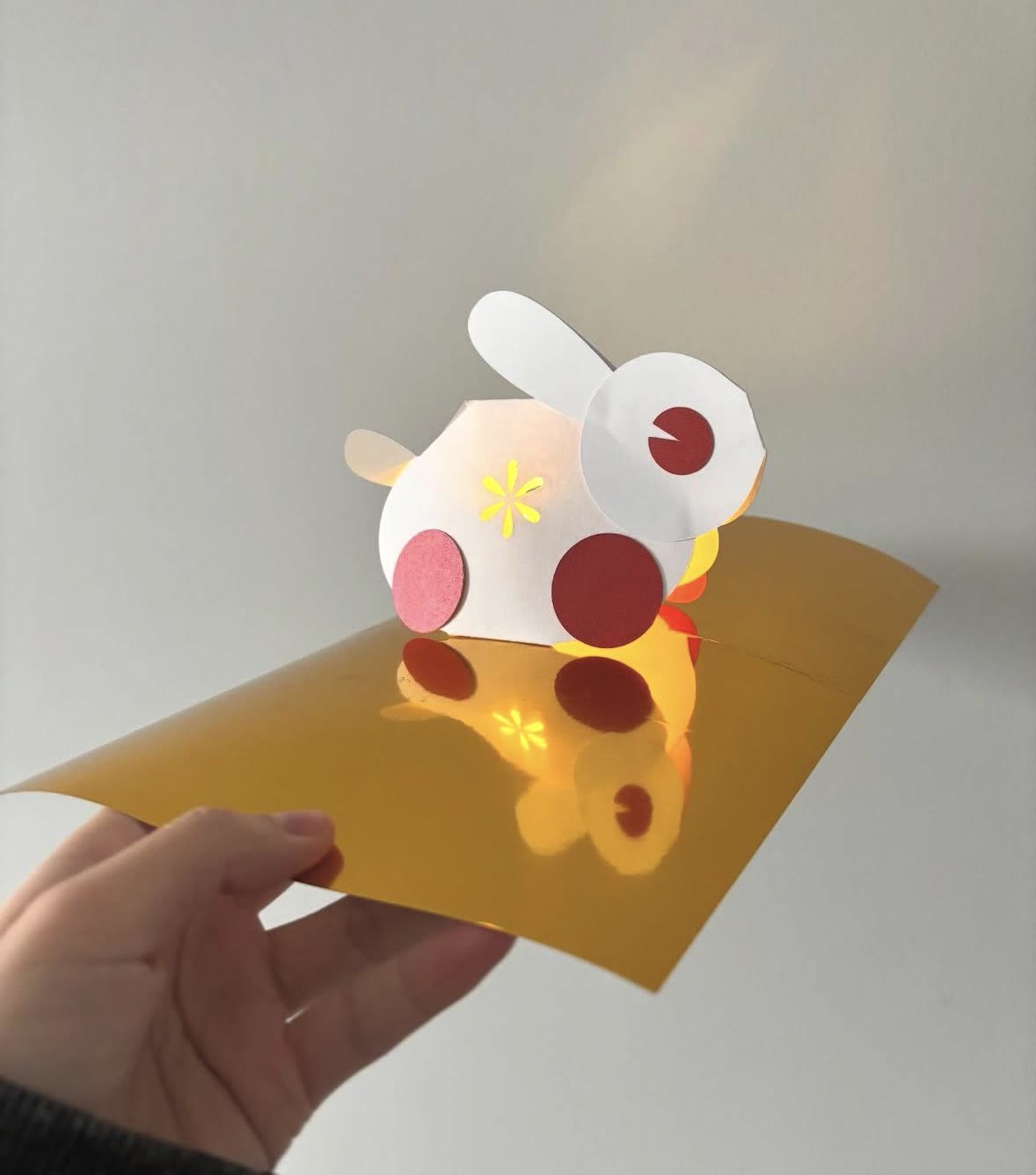 A white paper rabbit pop-up card with red cheeks on gold paper, lit from behind