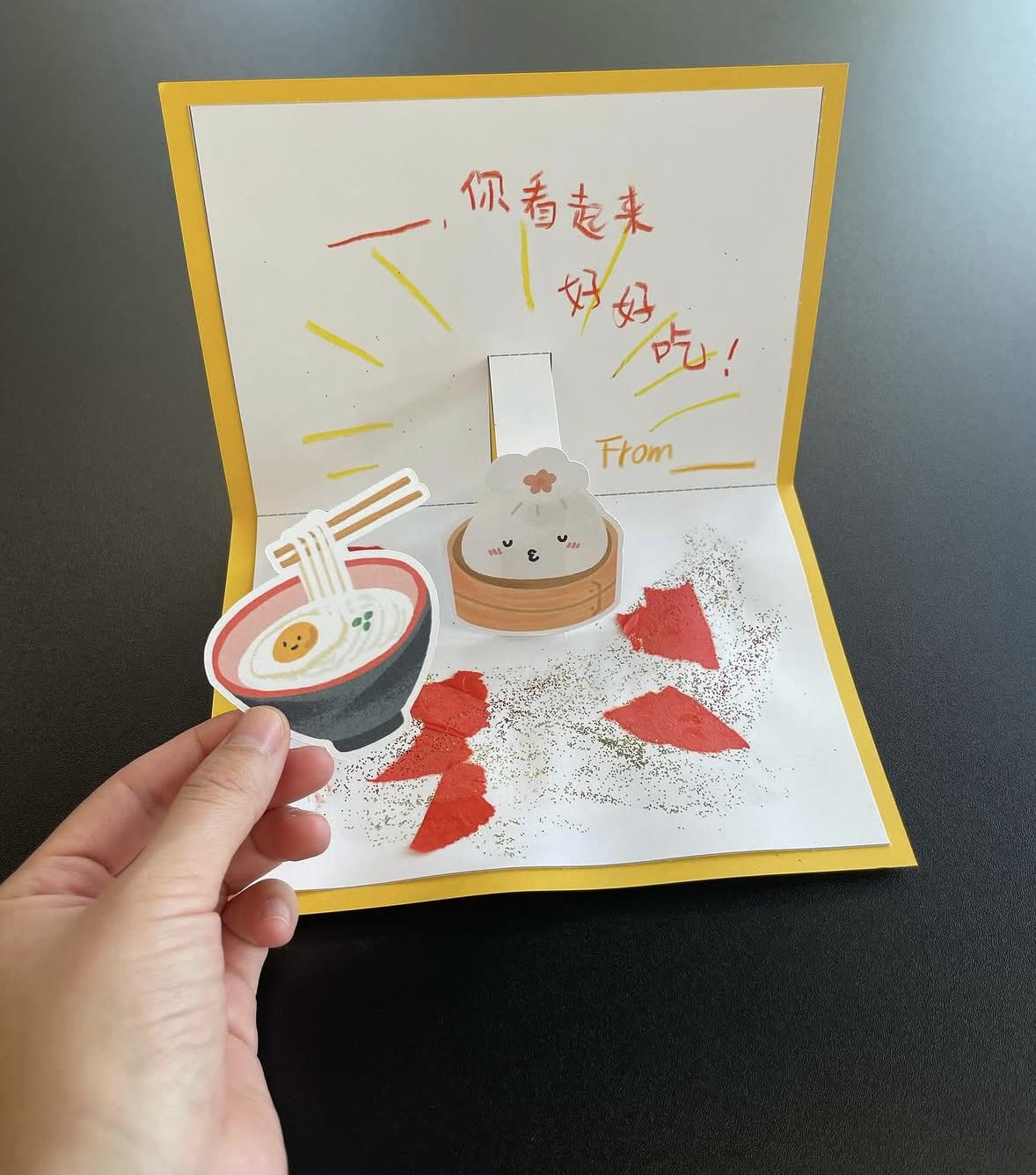 A food pop-up card: a bowl of noodles and a steamed bun standing up from a yellow card, with hand-written Chinese