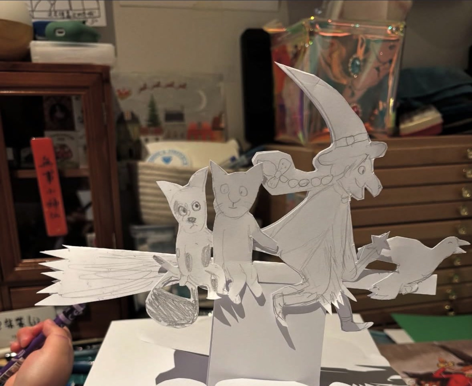 Making the card: the cut-paper witch, cat, cauldron and crow held up over a white cloud shape, with a pencil in hand