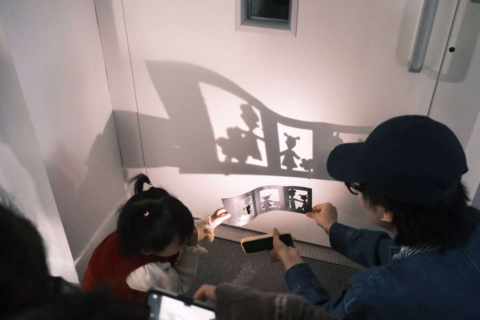 A workshop: a child and the artist projecting paper shadow shapes onto a white wall with a phone torch