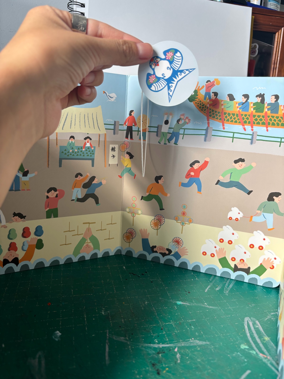 《百宝箱》 unfolded into a street panorama, with a hand holding a round cut-out